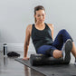 Woman using black SKLZ foam roller on leg while seated on exercise mat, with fitness ball in background