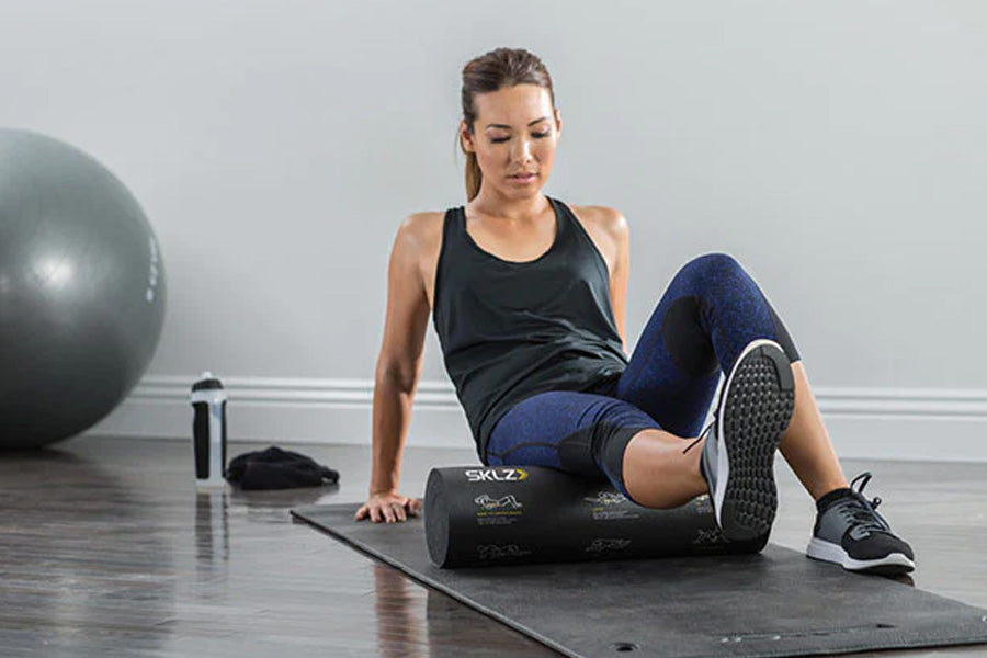Woman using black SKLZ foam roller on leg while seated on exercise mat, with fitness ball in background