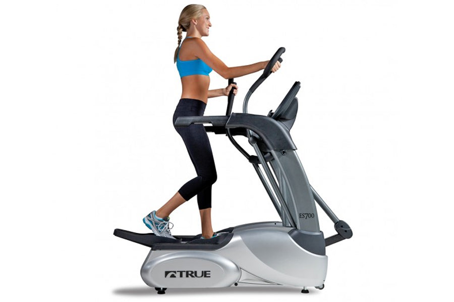 Woman in blue sports bra and black capris using TRUE ES700 elliptical machine with gray and white frame on white background