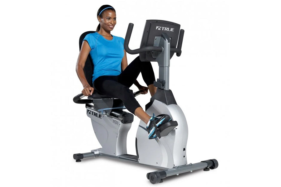 Woman in blue shirt and black leggings using white and black TRUE recumbent exercise bike with digital display on white backg