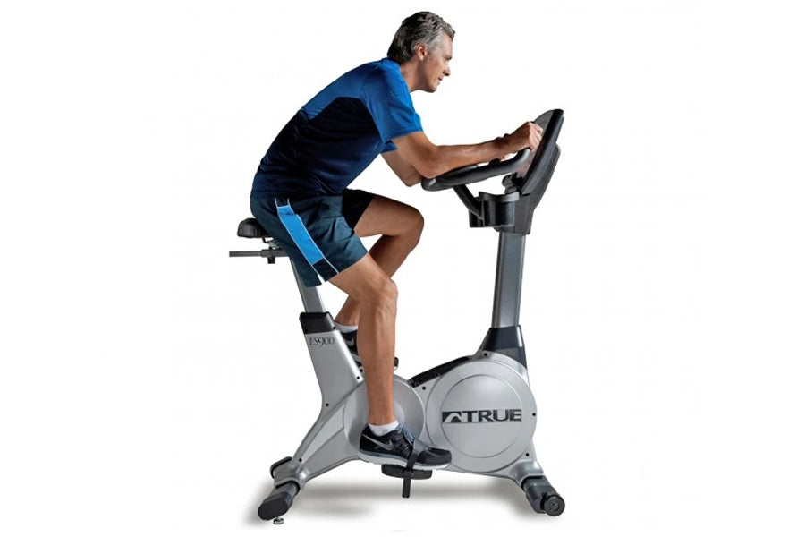 Man wearing blue and black athletic shirt and shorts riding gray TRUE upright exercise bike on white background