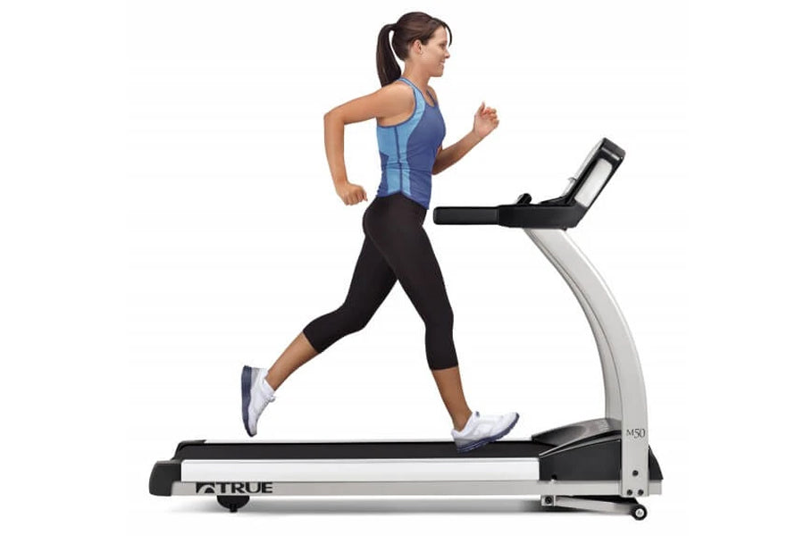 Woman running on TRUE M50 treadmill with blue tank top and black capri leggings, white sneakers, on white background