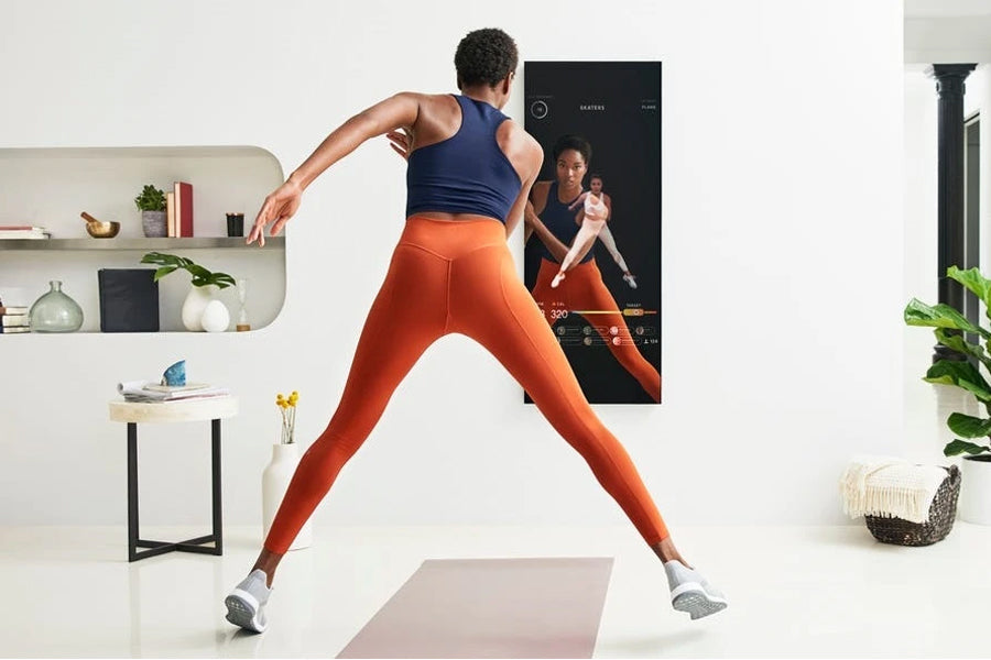 Person wearing navy tank top and orange leggings exercising in front of black Lululemon Studio Mirror on white wall