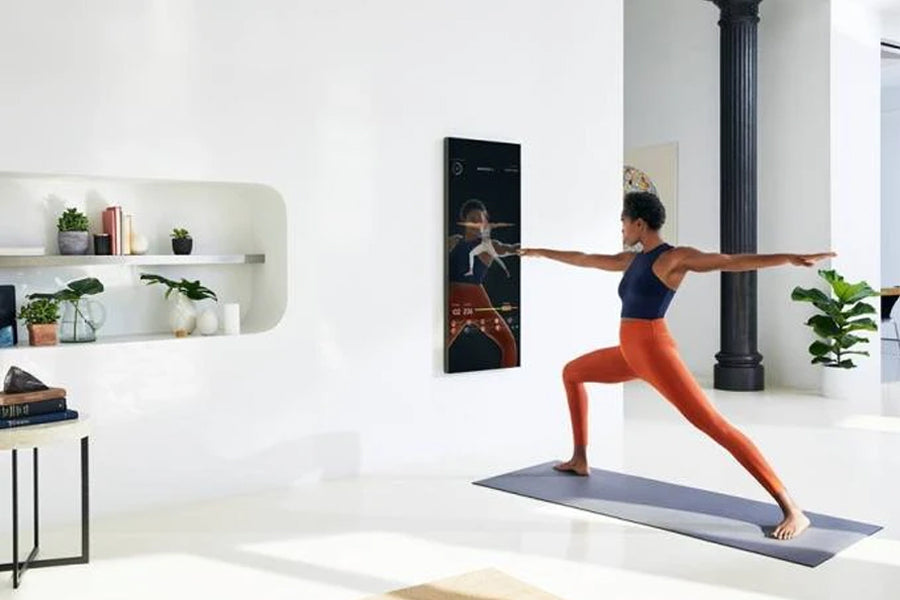 Woman in navy sports bra and orange leggings performing yoga pose on blue mat in front of Lululemon Studio Mirror mounted on 