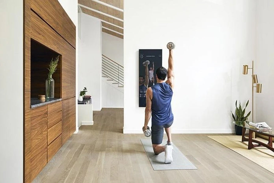 Man in blue tank top and black shorts using lululemon Studio Mirror Pro on white wall in home gym space
