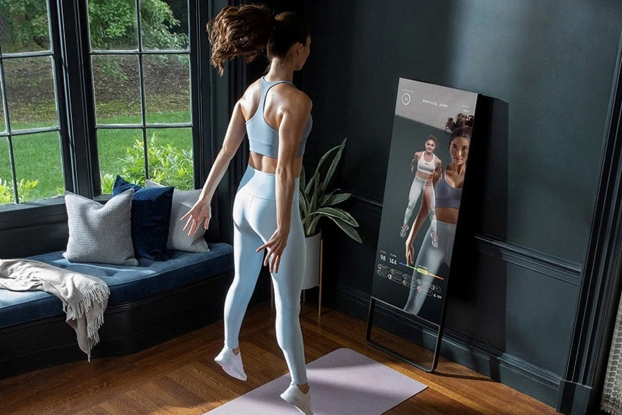 Lululemon Studio Mirror displayed in home gym setup, woman in white sports bra and leggings viewed from behind