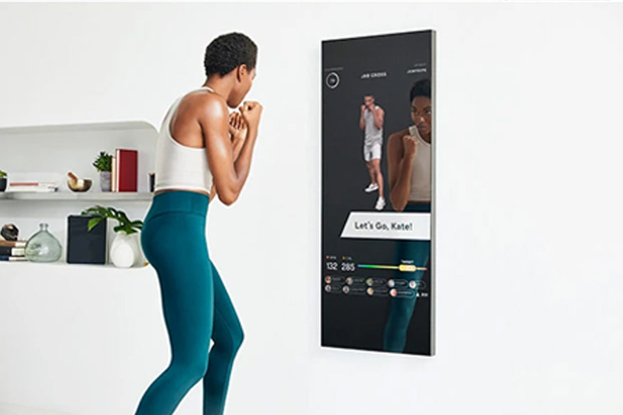 Woman in white tank top and teal leggings viewed from side using lululemon Studio Mirror Pro with fitness class displayed on 