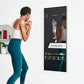 Woman in white tank top and teal leggings doing boxing workout in front of black interactive mirror displaying fitness class 