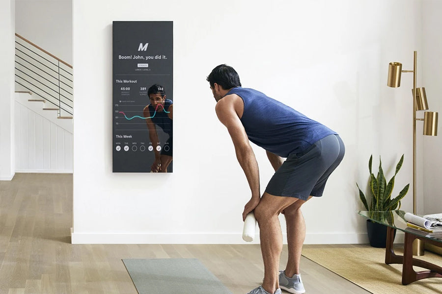 Man in blue tank top and black shorts performing exercise on gray mat in front of wall-mounted dark digital fitness mirror di