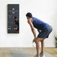 Man in blue tank top and black shorts doing a bent-over exercise pose on gray mat in front of wall-mounted black digital fitn