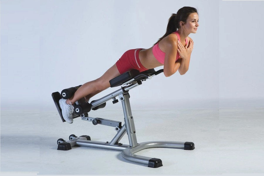 Woman in pink sports top and black shorts using silver hyper-extension bench with leg extension attachment on white backgroun