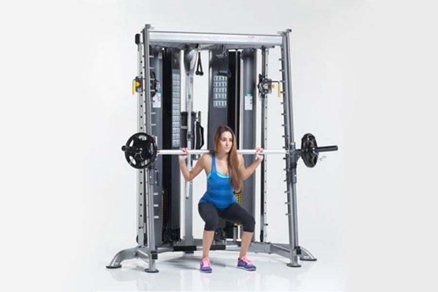 Woman in blue tank top performing squat exercise on gray and black Smith press machine with weighted barbell