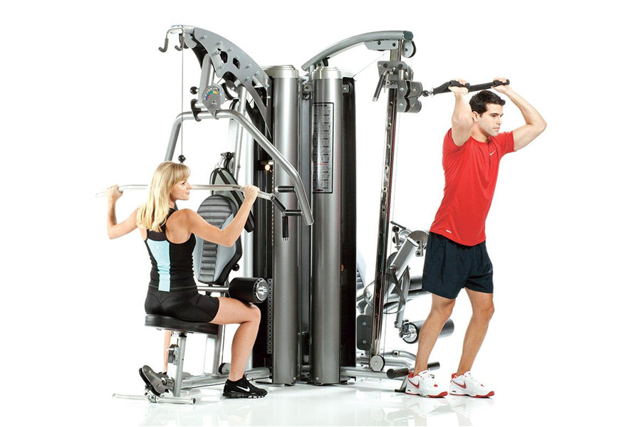 TuffStuff Apollo 7300 three-station multi-gym system with silver steel frame, two people demonstrating different exercises on