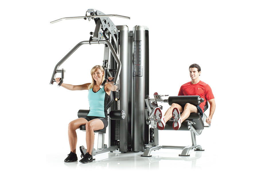 TuffStuff Apollo 7200 2-station multi gym system with woman using chest press and man using leg extension, shown on white bac