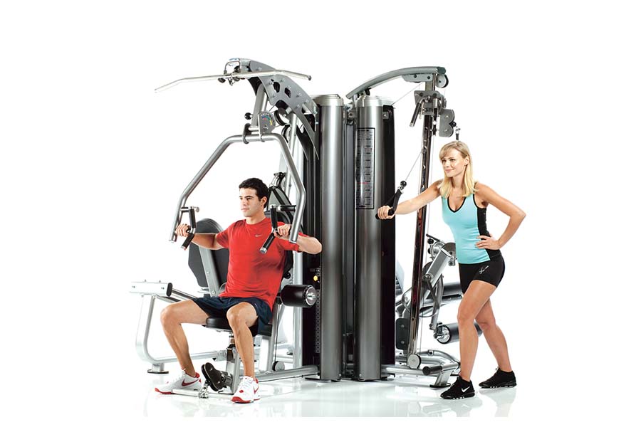 TuffStuff Apollo 7400 4-station multi-gym system with silver metal frame, shown with male model using chest press and female 
