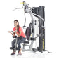 Woman in coral tank top and patterned leggings demonstrating TuffStuff Classic Home Gym with black and silver frame on white 