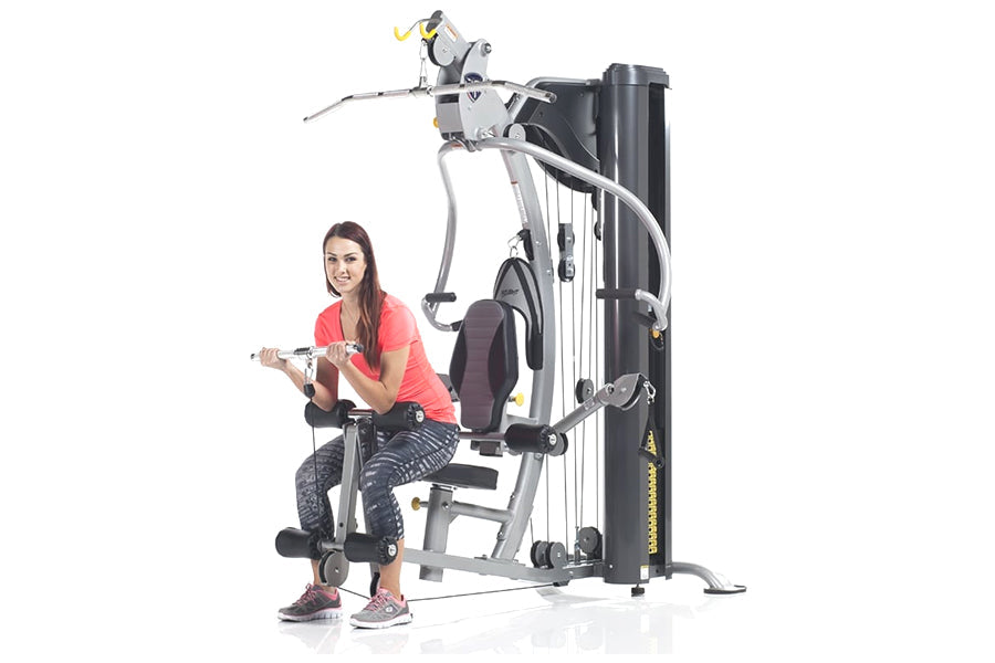Woman in coral tank top and patterned leggings demonstrating TuffStuff Classic Home Gym with black and silver frame on white 