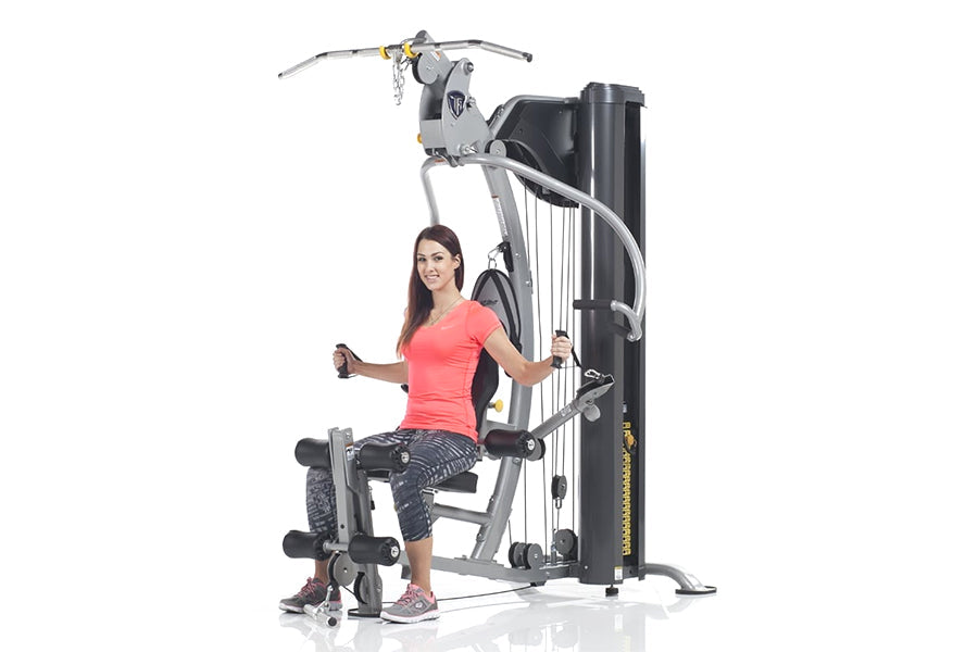 Woman in coral shirt and dark leggings using TuffStuff Classic Home Gym with gray and black frame on white background