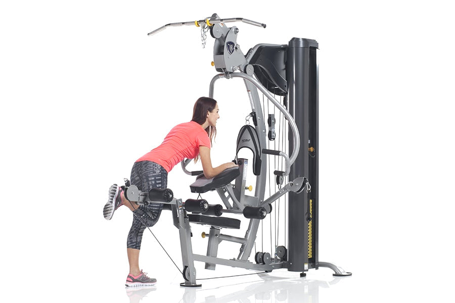 Woman in coral shirt and gray capris using silver and black multi-station home gym machine on white background