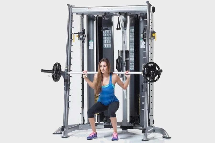 Woman in blue tank top and black capris performing squat exercise on silver and black multi-functional trainer home gym syste