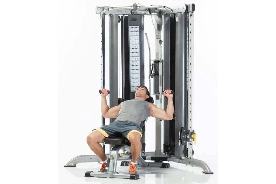 Man using silver and black multi-functional gym trainer with dual cable stations and smith press attachment, seated on bench 