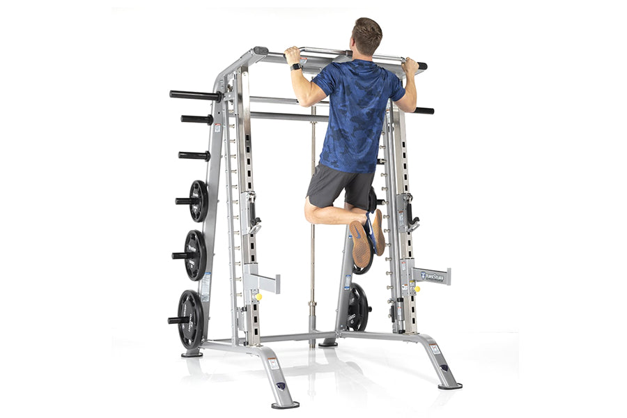 Man performing pull-up on silver and black TuffStuff Evolution Smith Machine with weight plate storage, worn by model on whit