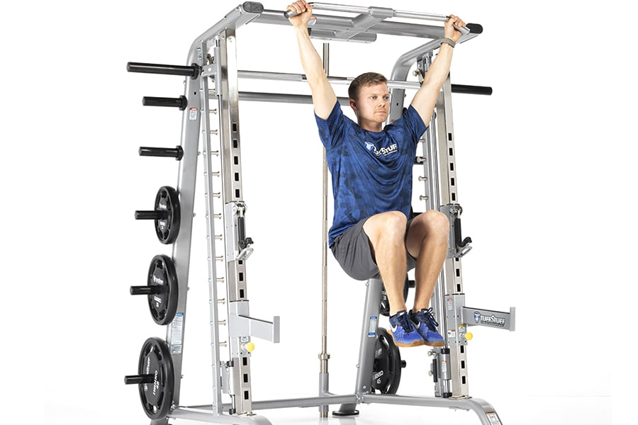 Man using silver TuffStuff Smith machine with black weight plates, performing pull-up exercise on horizontal bar