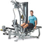 Gray and black multi-station home gym with male model in blue shirt demonstrating leg press position on white background