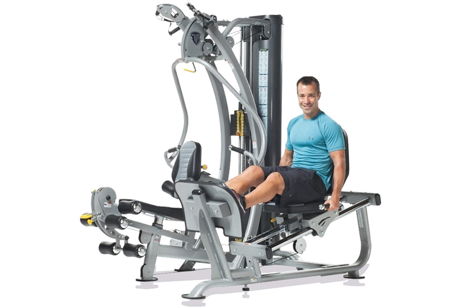 Gray and black multi-station home gym with male model in blue shirt demonstrating leg press position on white background