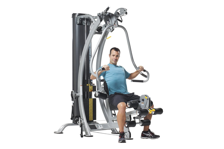 Man in blue shirt using silver and black multi-station home gym machine with leg press attachment, on white background
