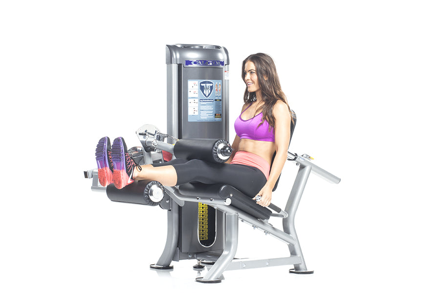 Woman in purple sports bra and pink shorts using black and silver leg extension machine with weight stack on white background