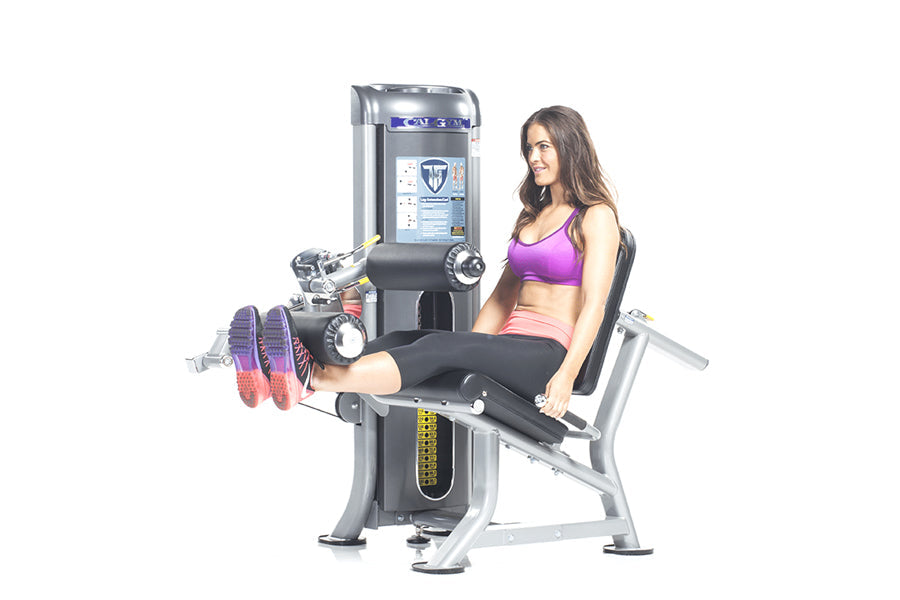 Woman using black and silver leg extension machine with purple sports bra and pink shorts on white background