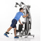 Male athlete using silver and black functional trainer machine with dual cable pulleys, wearing blue shirt and navy shorts, o