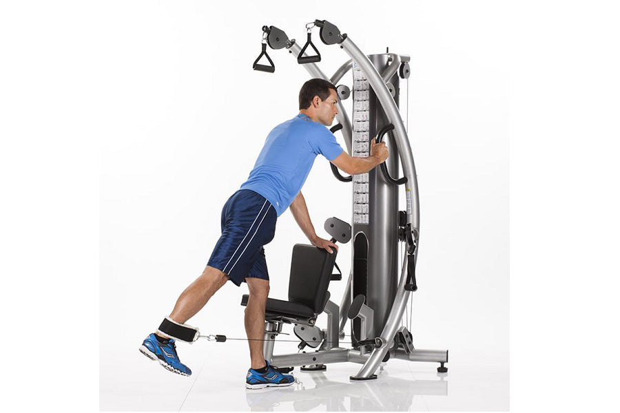 Male athlete using silver and black functional trainer machine with dual cable pulleys, wearing blue shirt and navy shorts, o