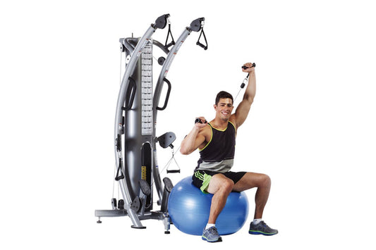 Man performing shoulder exercise on silver TuffStuff functional trainer with dual pulleys, seated on blue stability ball