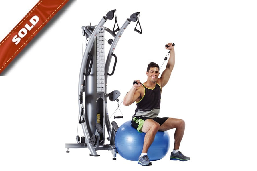 Man demonstrating gray and black functional trainer machine with dual pulleys, seated on blue stability ball with arms raised