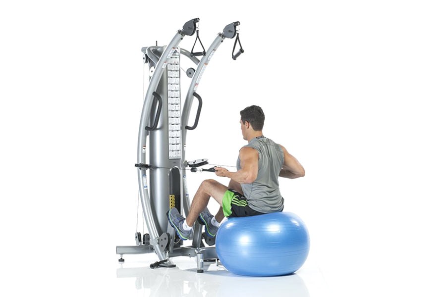 Man using silver functional trainer machine with dual cable arms, seated on blue stability ball, on white background