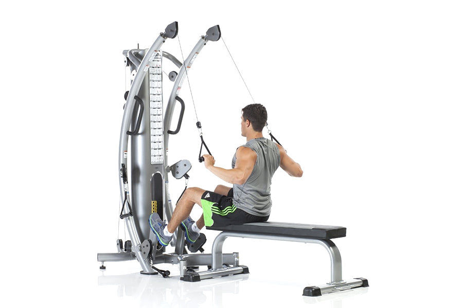 Male user demonstrating silver and black cable machine trainer with dual pulleys and height measurement scale, seated on blac