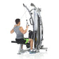 Man using gray and black functional trainer machine with dual cable system, seated on black bench pad, on white background