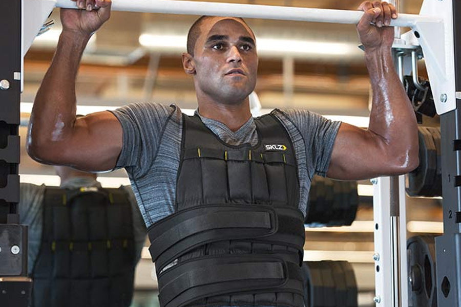 Man wearing SKLZ black weighted vest with gray athletic shirt, performing pull-up exercise in gym