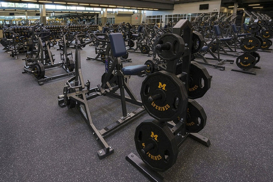 Busy gym floor with stationary bikes, weight plates, and dumbbells on gray interlocking rubber tiles