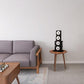 Black four-speaker stacked tower with white cone drivers on wood side table in modern living room setting