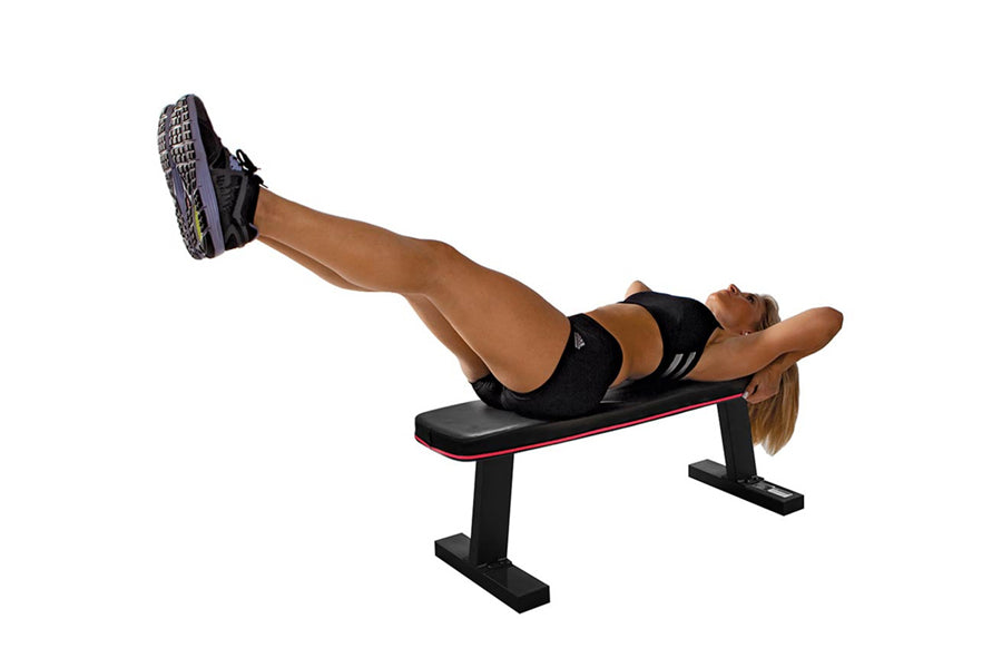Woman performing leg exercise on black flat weight bench with pink trim, wearing black sports bra and shorts with gray athlet