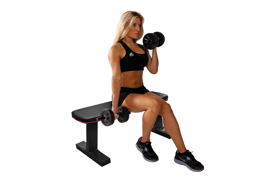 Woman in black sports bra and shorts sitting on black flat weight bench, holding dumbbell in bicep curl position, white backg