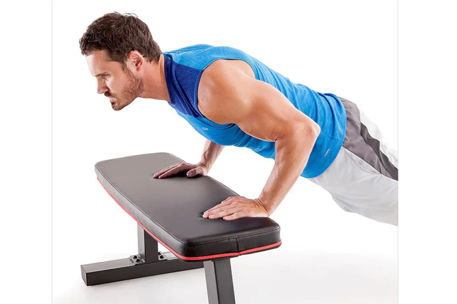 Man in blue tank top performing push-up exercise on black and red flat weight bench against white background