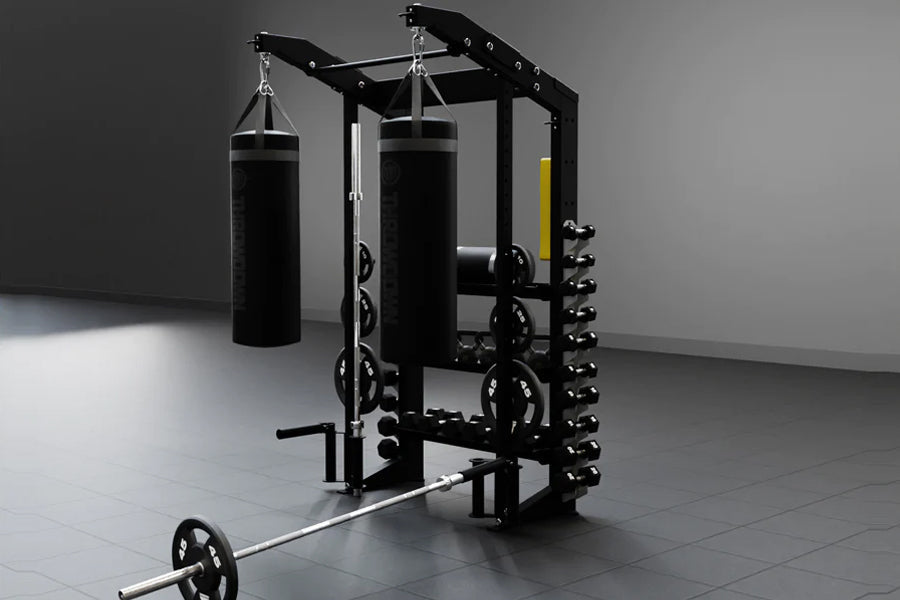 Black steel functional strength station with dual heavy bags, lat pulldown, plate storage, and barbell on gray floor