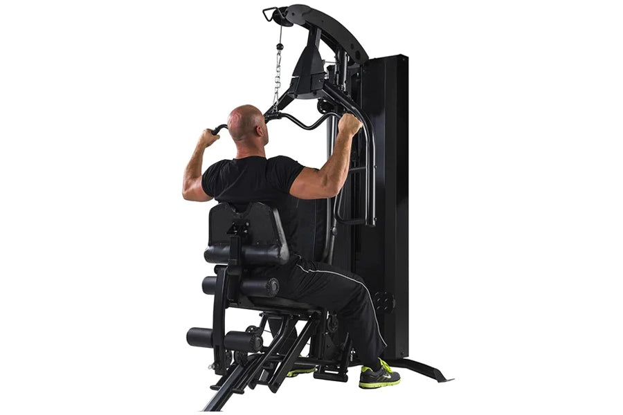 Black multi-station home gym with leg press, man in black shirt demonstrating lat pulldown exercise on white background