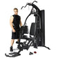 Man in black sleeveless shirt standing next to black multi-station home gym equipment with leg press and padded seat
