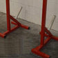 Pair of red heavy-duty squat stands with silver adjustable safety bars on concrete floor against white brick wall