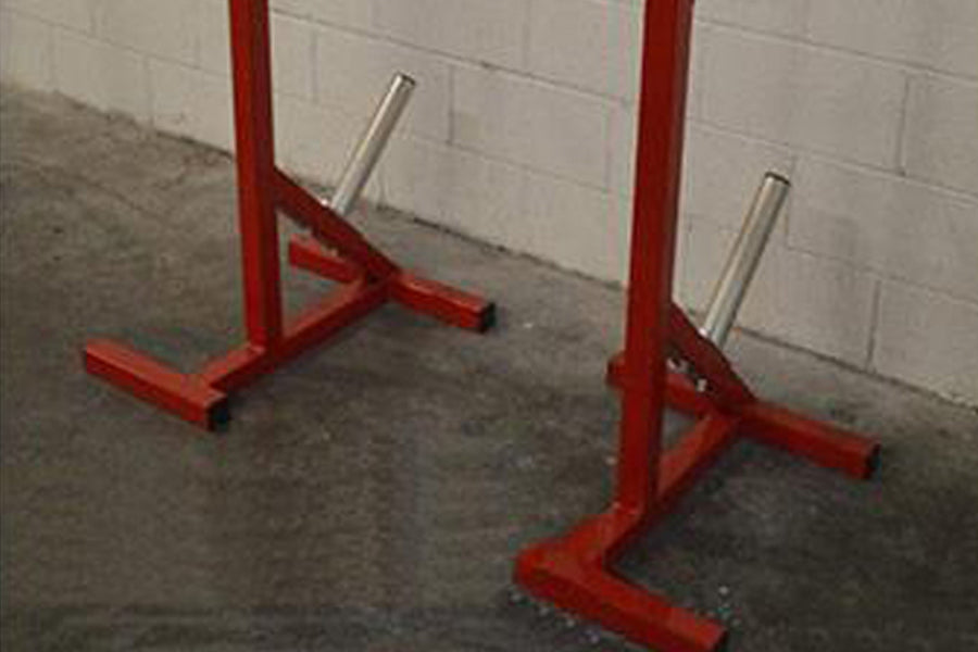 Pair of red heavy-duty squat stands with silver adjustable safety bars on concrete floor against white brick wall